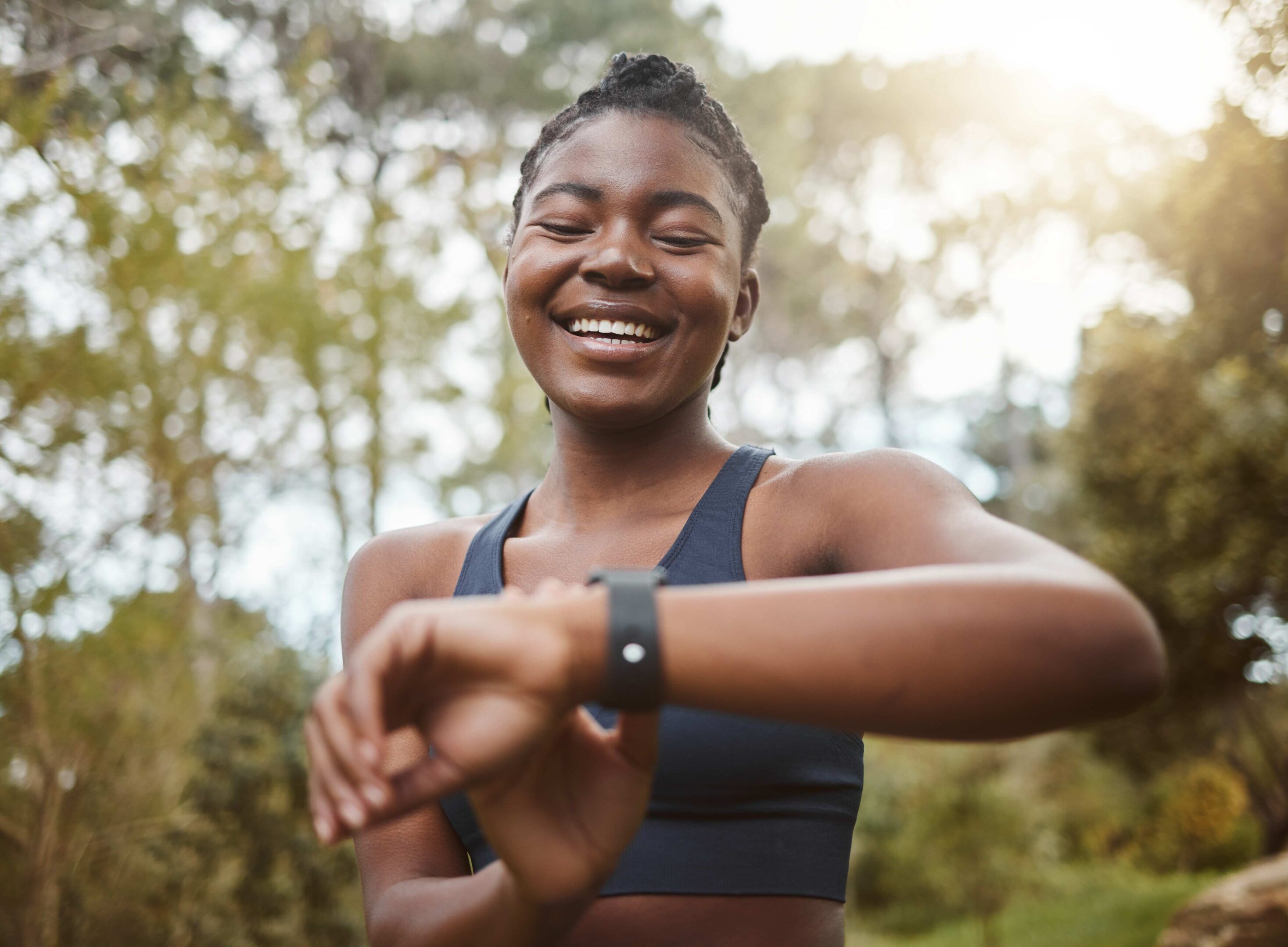 Woman in athletic wear smiles while looking at a smartwatch outdoors, with trees in the background.