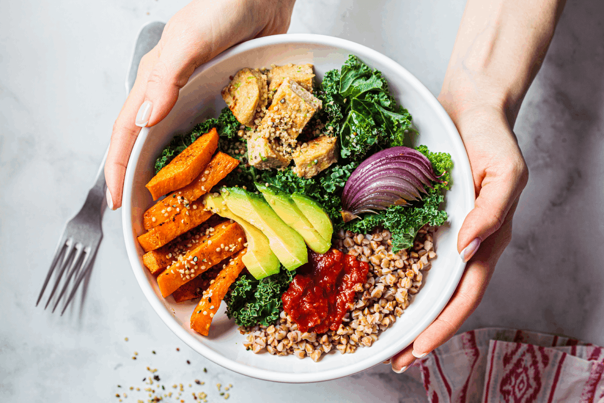 A bowl with sliced avocado, cooked sweet potatoes, red onion, greens, salsa, and seasoned grains, held by two hands. A fork is visible on the left side.